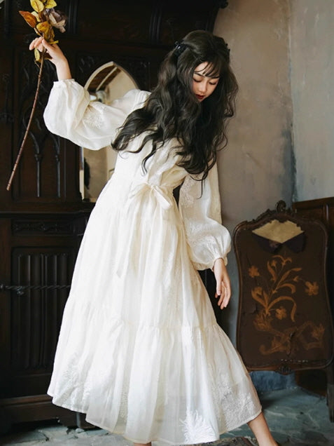 A woman in an elegant white fantasy dress with puff sleeves and a tiered skirt, posing delicately against a vintage chair and ornate wooden cabinet.