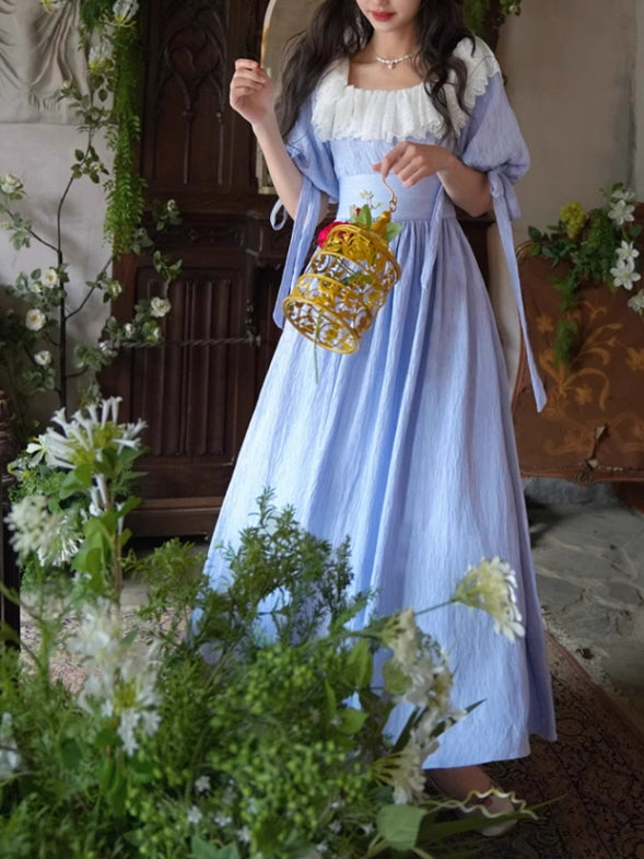 A fairytale-inspired vintage blue dress with a delicate white lace collar, styled with puffed sleeves tied with ribbons, creating a soft fantasy aesthetic among lush greenery.