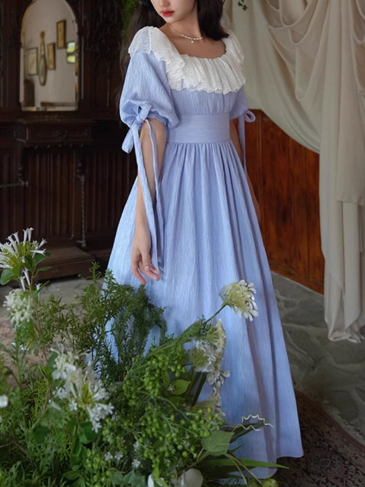 A model wearing a fairytale-inspired vintage blue dress with a lace-trimmed neckline and tied puff sleeves, standing gracefully among lush greenery.