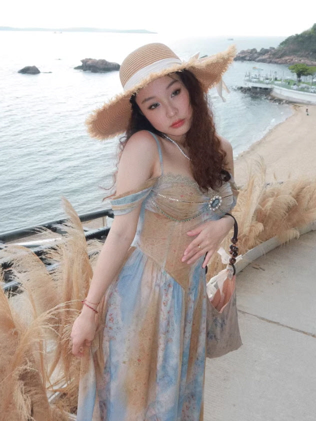 A woman in a vintage-inspired, blue and gold off-shoulder gown adorned with lace and jeweled accents, posing by the seaside railing with a romantic, dreamy gaze.