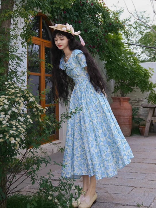 A vintage-inspired blue floral dress with a ruffled collar and flowing skirt, styled with a charming straw hat for a dreamy cottagecore vibe.