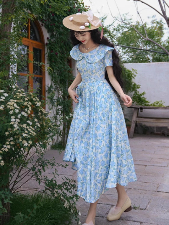 A graceful blue floral gown with a flowing skirt and peter pan collar, styled with a whimsical straw hat adorned with floral accents.