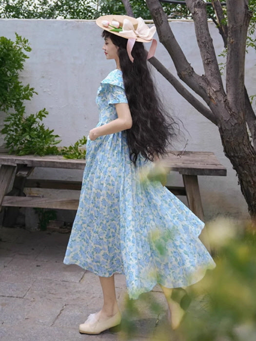Side view of a flowing blue floral dress with a wide collar and puff sleeves, creating a dreamy, vintage-inspired silhouette under a garden tree.