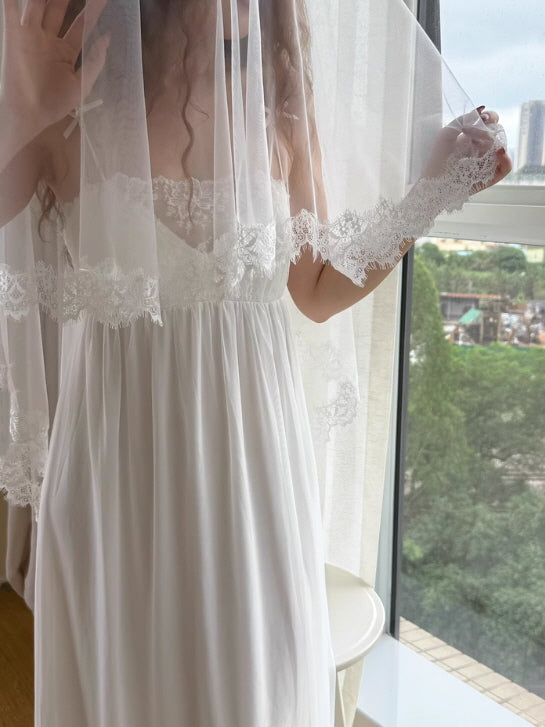 A woman in a white lace-trimmed nightgown holds a sheer veil over her face, creating a dreamy, romantic fairycore moment by the window.