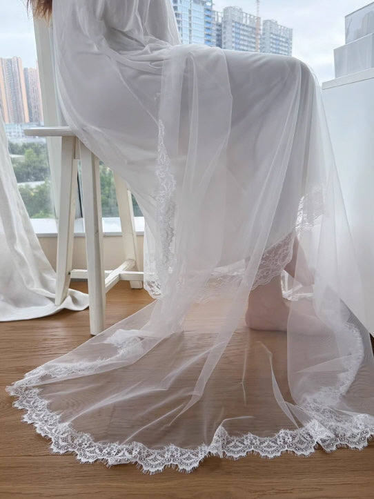 Soft tulle and lace hem drape elegantly on the floor as a woman in a white vintage nightgown sits by the window in a serene princesscore scene.