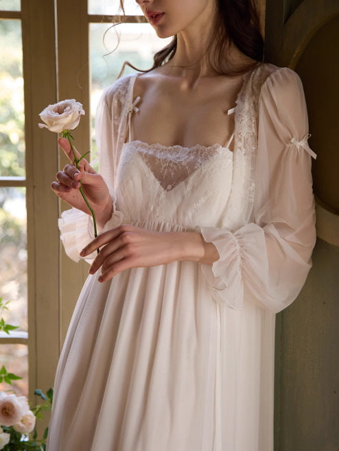 A woman in a sheer white lace nightgown gently holds a single blush rose by the window, surrounded by soft sunlight and romantic calm.