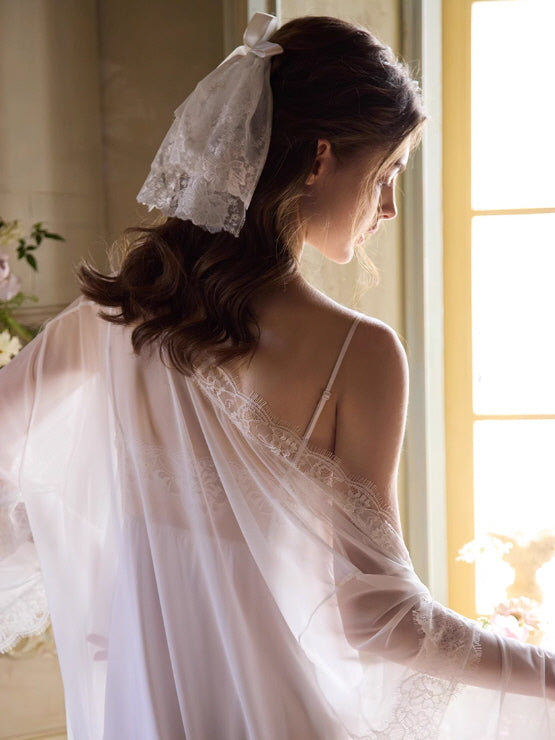A woman with soft curls and a lace ribbon in her hair wears a sheer white robe with floral lace trim, standing by a sunlit window in a vintage room.