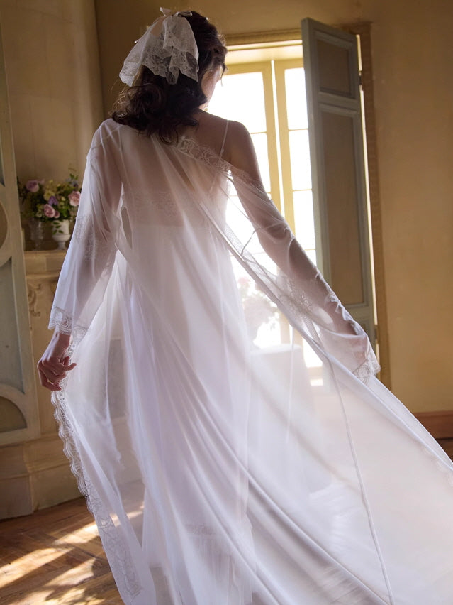 Captured from behind, the woman walks gracefully in a flowing sheer white robe, the delicate lace hem catching light in a romantic vintage interior.