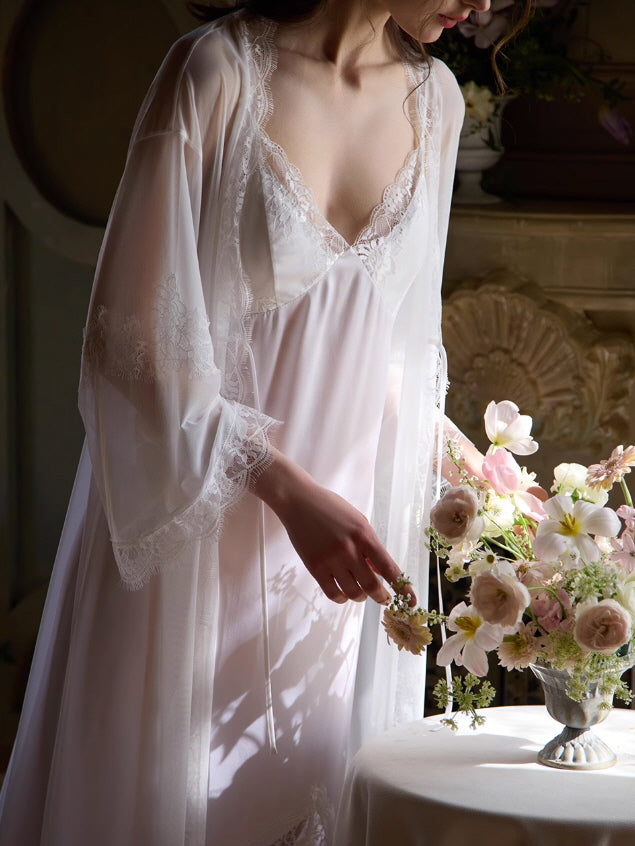Close-up of a feminine hand delicately reaching for pink blossoms, wearing a sheer fairycore robe with intricate lace trim in a romantic morning light.