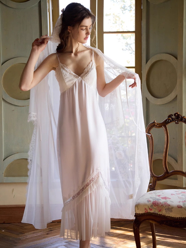 A model in a vintage-inspired ivory slip dress with a sheer lace robe lifts the fabric gracefully by a sunlit window, evoking a fairytale aesthetic.