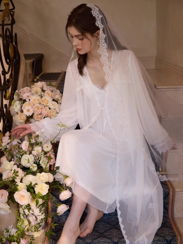 A model sits gracefully on stairs surrounded by roses, wearing a romantic white loungewear gown with lace trim and a sheer robe in fairycore style.