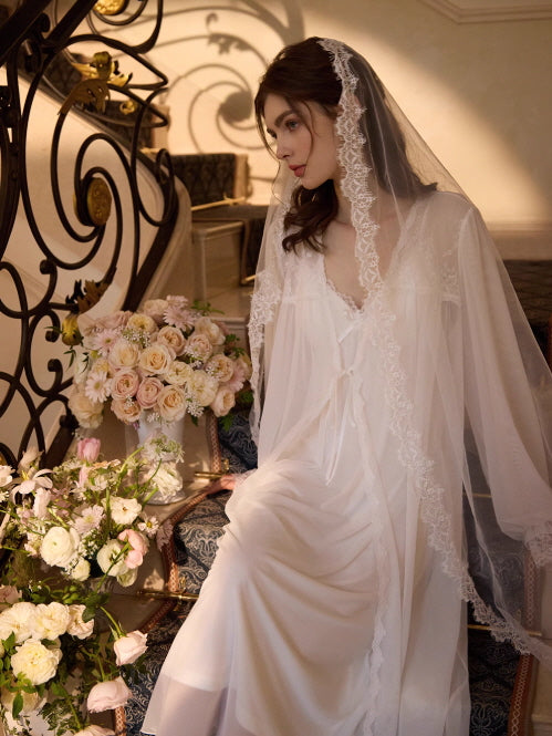 Seated by cascading flowers, the model gazes softly in a vintage-inspired white loungewear set with lace details and a delicate layered silhouette.