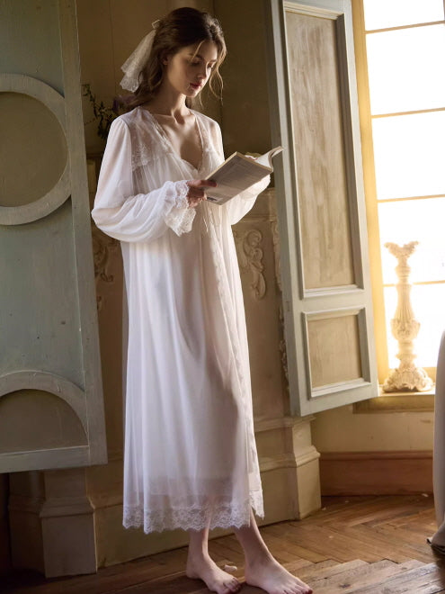 A woman in a romantic white loungewear gown with sheer long sleeves and lace edges stands by the window, reading a book in soft morning light.