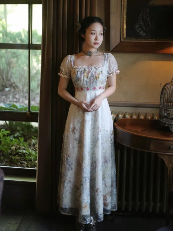 Full-body view of a Romantic Fairytale vintage-inspired floral lace maxi dress featuring a square neck, short puff sleeves, embroidered waist band, and flowing skirt, styled indoors by a window for a fairycore vintage aesthetic outfit.