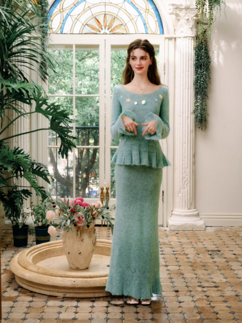 A romantic mint green knit top styled with a matching long skirt, featuring a fitted waist, flared peplum detail, and elegant long sleeves, captured in a bright vintage interior with botanical surroundings.
