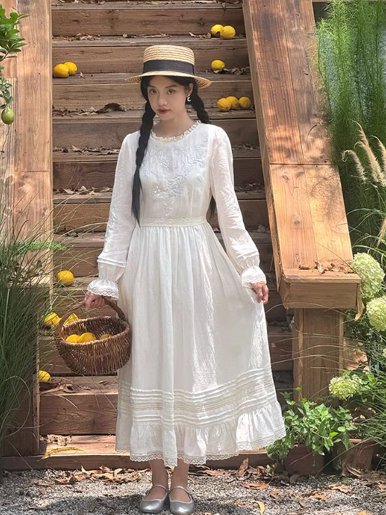 A woman wearing a white embroidered long sleeve dress with lace trim, styled in a cottage garden setting, featuring a flowing vintage silhouette and romantic cottagecore aesthetic outfit.