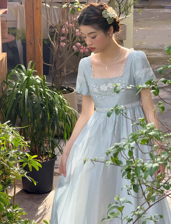 Ethereal woman with closed eyes wearing a dreamy pale blue fairytale gown with puff sleeves, posing in a sunlit garden for a romantic princesscore aesthetic.