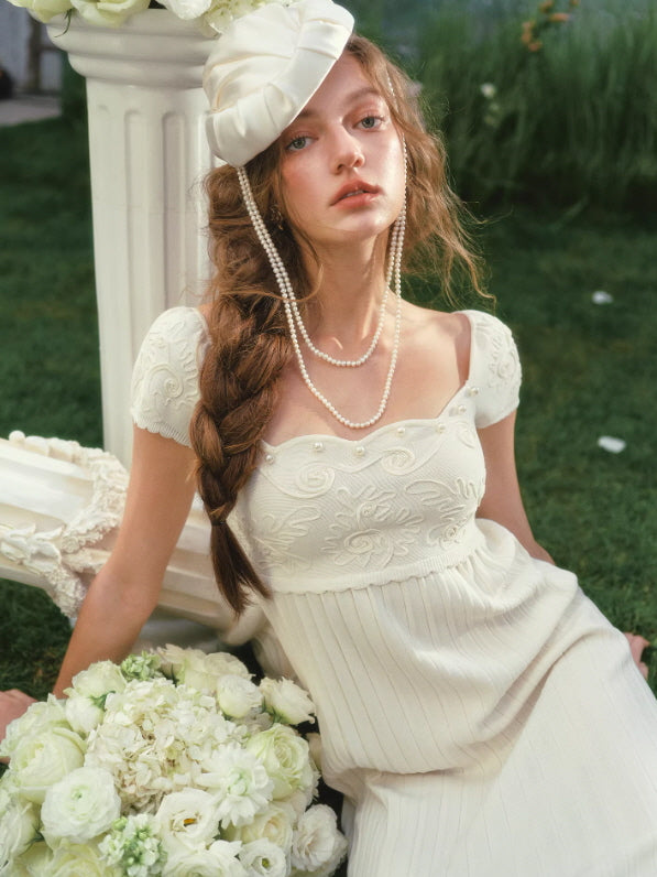 Elegant ivory cottagecore dress with floral embroidery bodice and pleated skirt, model seated on grass with white roses