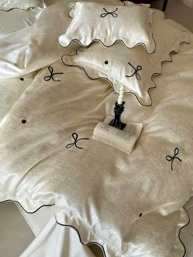 Romantic ivory bedding with delicate black ribbon bow details, wavy piping, and lustrous satin texture