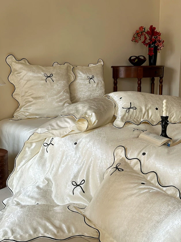 Princesscore ivory bedding with glossy satin fabric, black bow accents, and softly ruffled scalloped edges