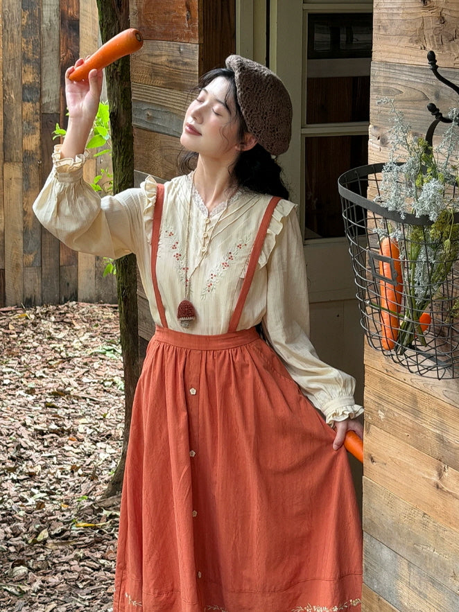Vintage prairie style outfit with cream ruffle blouse and terracotta suspender skirt beside a wooden cottage