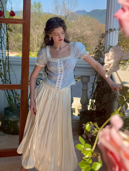 A model wearing a pastel blue corset top with lace accents and puff sleeves, paired with a flowing cream midi skirt, creating a dreamy, fairytale look amidst a romantic garden setting with blooming roses.