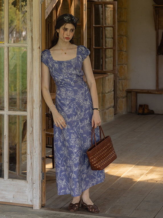 Woman in a blue floral puff-sleeve dress holding a woven bag, standing on a rustic wooden porch, capturing a vintage cottagecore aesthetic.