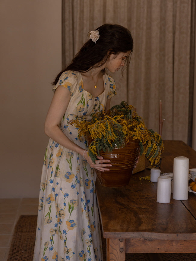A young woman with wavy hair gently adjusts a large pot of vibrant yellow flowers on a rustic wooden table. She wears a cream floral dress adorned with blue and yellow blossoms, exuding a vintage, cottagecore ambiance in a softly lit room.