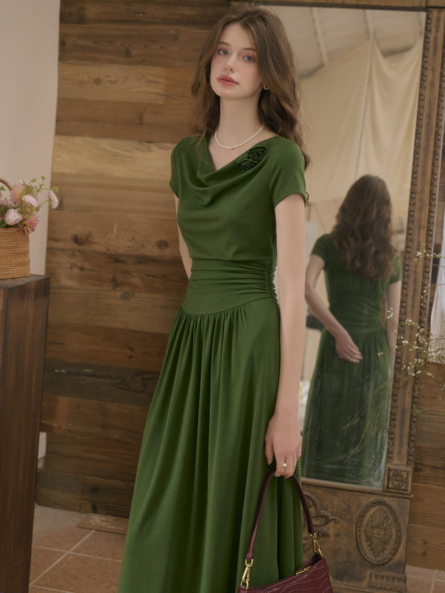Model in a soft green dress with a cinched waist and delicate flower brooch detail, standing gracefully in front of a large ornate mirror. Romantic indoor scene with warm vintage decor.