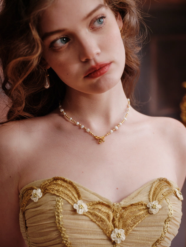 Detailed close-up of a gold floral corset with pearl necklace, capturing the model’s soft expression and romantic curls in a vintage-inspired fairytale setting.