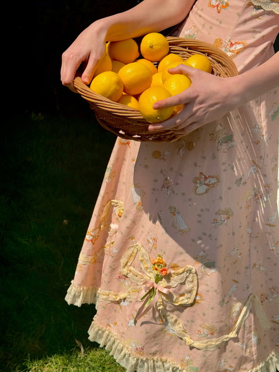 Detailed close-up of a whimsical floral dress adorned with lace and bow accents, as a wicker basket of lemons adds a charming, cottagecore touch.