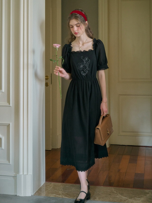 A young woman in a retro black vintage dress with floral embroidery stands indoors holding a pink rose, wearing a red headband and classic leather handbag.