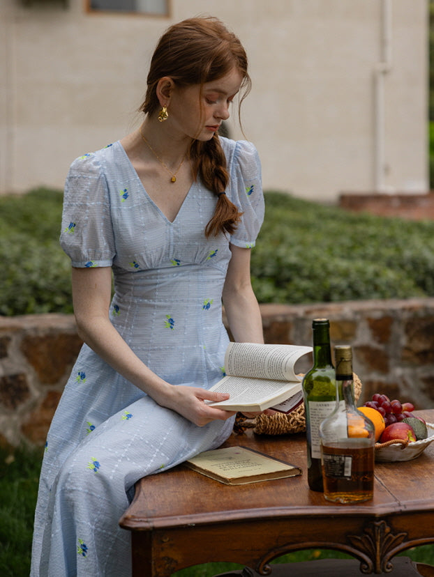 A woman in a blue floral midi dress with delicate puff sleeves sits at an outdoor wooden table, reading a book. Bottles, fruit, and a woven basket add a cozy, countryside ambiance to the scene.