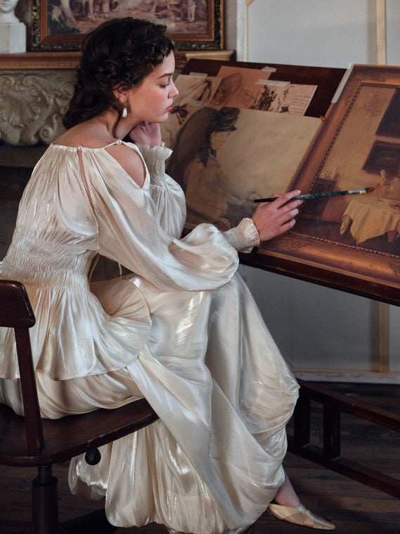 Seated at a wooden easel, a woman wearing a romantic ivory loungewear set delicately paints a classical scene, her off-shoulder blouse and flowing skirt reflecting a dreamy, vintage cottagecore mood.