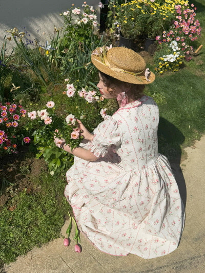 Seated by a flower bed, a woman in a puff-sleeved vintage cotton dress and straw hat gently reaches for pink blooms, evoking a soft and fairytale-like countryside mood in timeless women’s fashion.