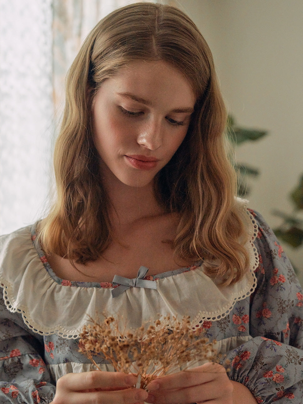 A serene moment as the model gently holds dried flowers while wearing a blue floral nightgown adorned with lace ruffles and a dainty satin bow.