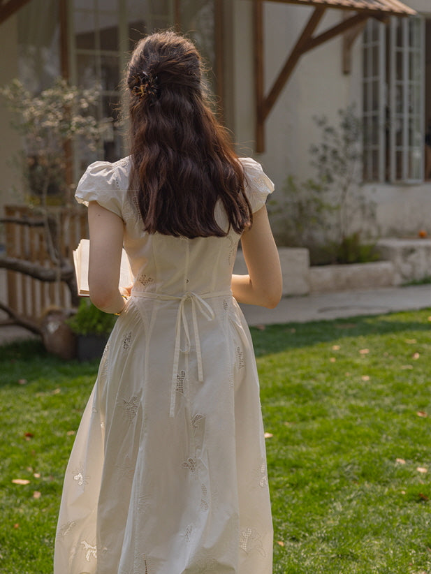 A woman standing on a green lawn, wearing a white embroidered long dress with a tie-back design, holding a book, and her long wavy hair styled with a hair clip.