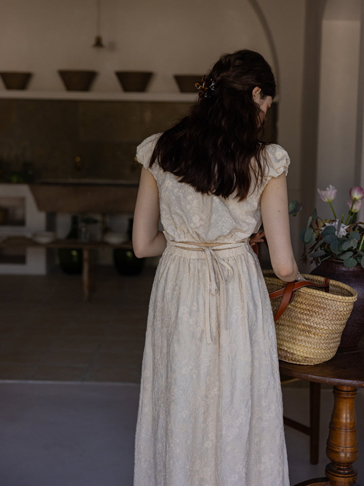 Back view of a woman in a cream-colored textured midi dress with a tied waist and puff sleeves, her hair styled in loose waves with a hair clip. A woven basket sits on a wooden table nearby, creating a cottagecore aesthetic.