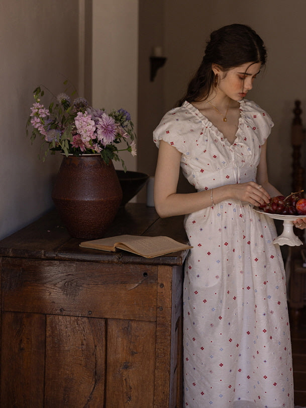 Young woman in a cream floral print dress with a ruffled neckline, holding a plate of grapes while standing beside a wooden cabinet with an open book and a rustic vase filled with flowers, evoking a nostalgic cottagecore ambiance.