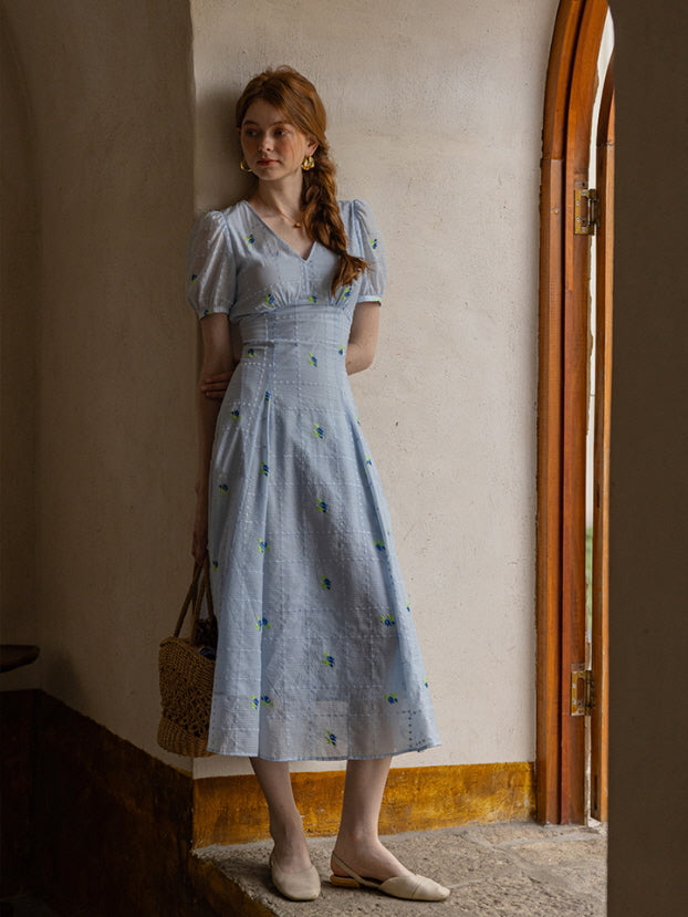 A woman stands against a rustic archway, wearing a light blue floral midi dress with puff sleeves and subtle embroidered details. She holds a woven basket, exuding a serene and vintage-inspired aesthetic.