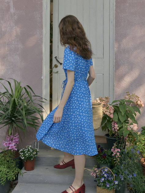 Woman walking gracefully in a blue floral vintage dress, captured from behind in a garden entrance filled with blooming flowers and soft shadows. Elegant and romantic outdoor moment.