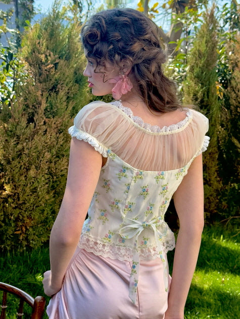 A back view of a floral corset top featuring sheer puff sleeves and lace trim, tied with a delicate ribbon at the waist, creating a soft, vintage-inspired silhouette in a garden setting.