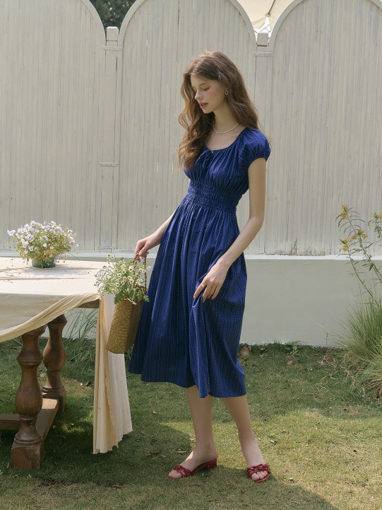 Woman in a navy blue smocked dress holding a basket of flowers, walking gracefully across a garden beside a rustic outdoor table. Soft fairytale charm with vintage summer elegance.