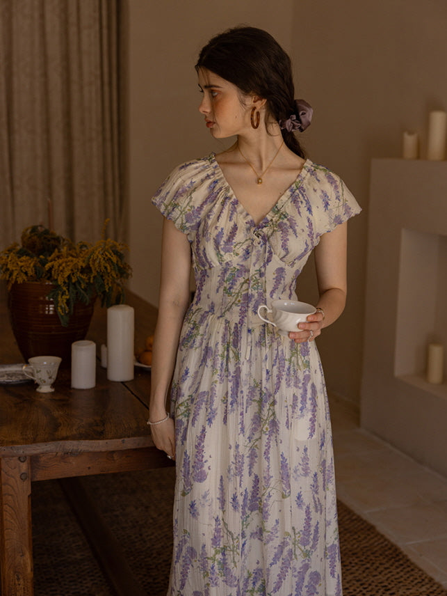 Woman in a lavender floral dress holding a teacup, standing by a rustic wooden table adorned with dried flowers and candles, creating a serene cottagecore atmosphere.