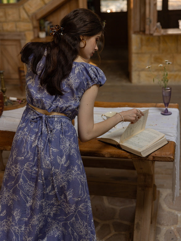 Back view of a woman in a blue floral cut-out dress with a tie-back detail, leaning over a rustic wooden table while flipping through a vintage book in a cozy cottage setting.