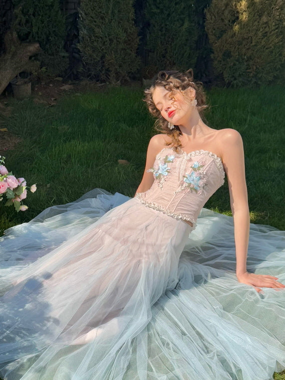 Romantic pastel corset dress with embroidered flowers and flowing tulle skirt, worn by a woman reclining on grass in sunlight – dreamy vintage fairytale fashion
