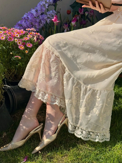 Close-up of ivory lace embroidered skirt with ruffle hem, styled with elegant heels and spring flowers in a garden – feminine romantic aesthetic outfit
