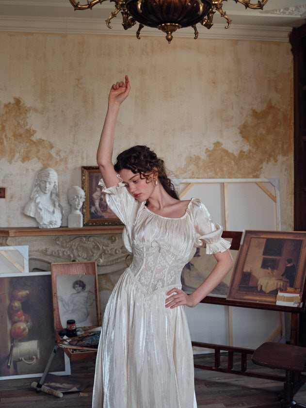 A woman posing gracefully in a softly shimmering ivory dress with puff sleeves and a lace corset, surrounded by vintage paintings and sculptures in an artist’s studio.