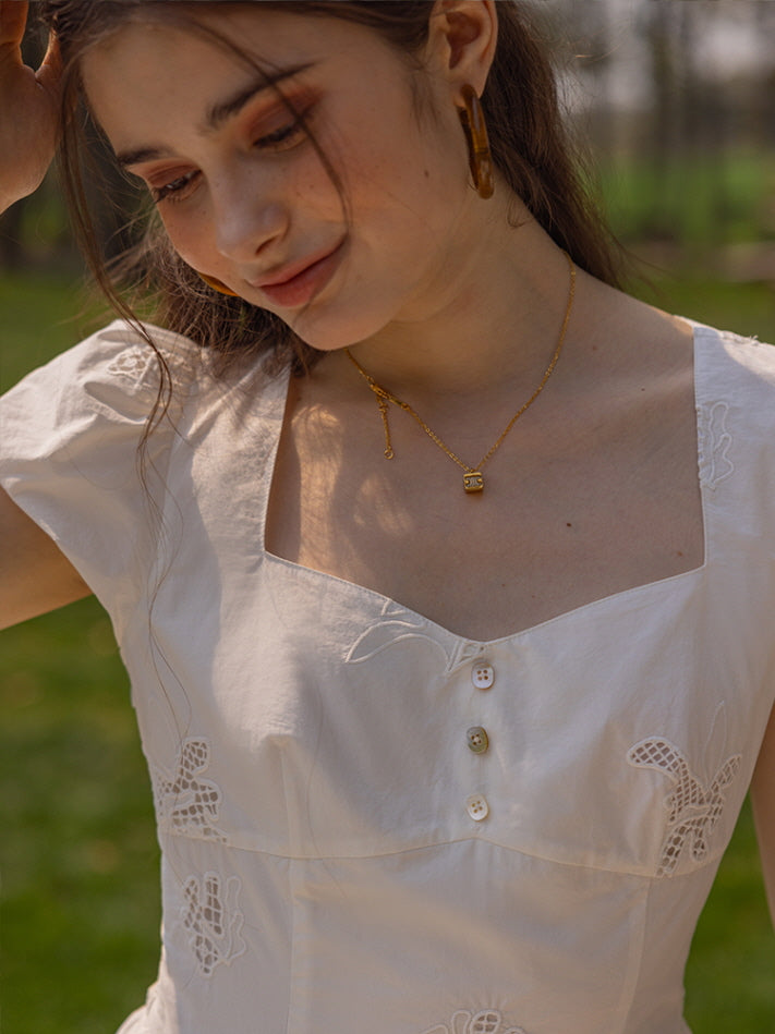 Close-up of a young woman smiling gently, showcasing the delicate square neckline and button-down bodice of a white embroidered dress, accessorized with gold hoop earrings and a dainty gold necklace.