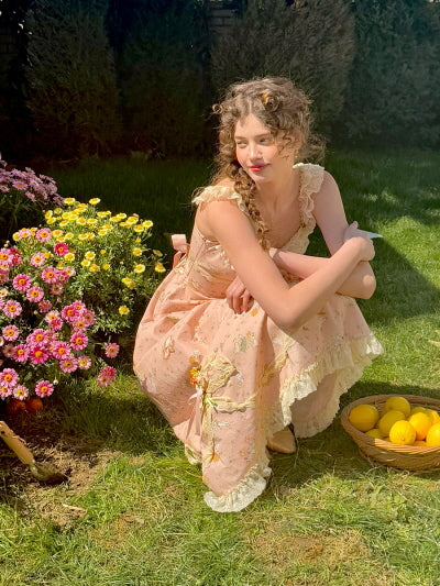 A romantic floral lace dress with delicate ruffle straps, as a young woman crouches beside vibrant garden flowers and a basket of lemons, evoking a fairytale picnic scene.